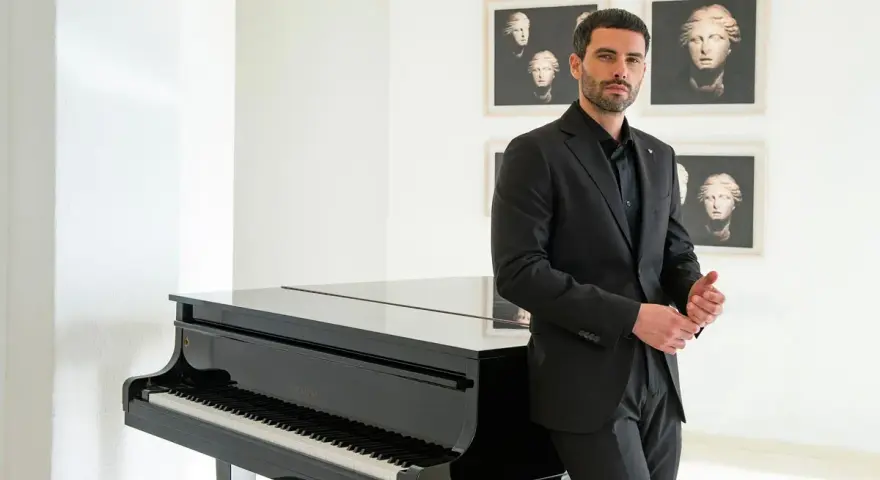 Uomo con elegante outfit per andare a teatro, in completo nero, accanto a un pianoforte a coda.