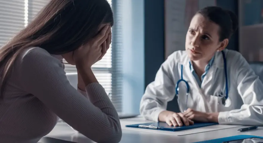 Kobieta w gabinecie lekarskim, ze smutkiem ukrytym w dłoniach, słucha diagnozy. Lekarka z troską pochyla się nad dokumentami, być może omawiając poronienie chybione.