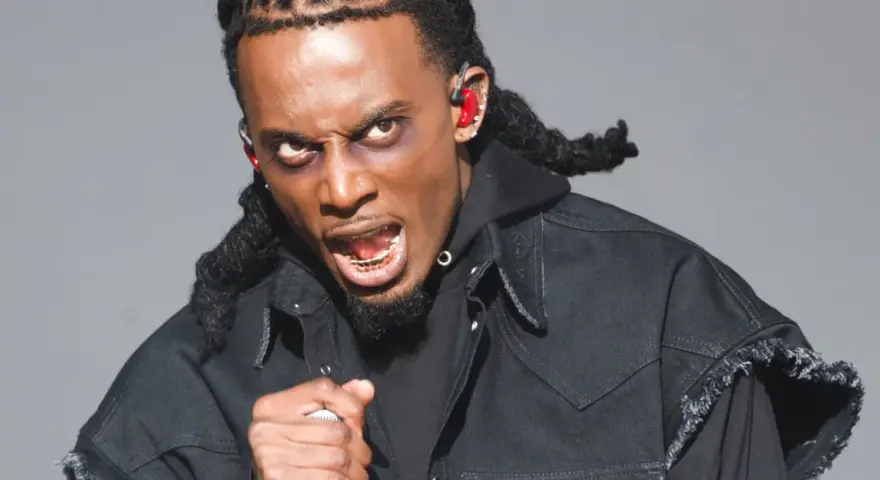 Playboi Carti, wiek nieznany, w czarnej kurtce z frędzlami, z mikrofonem w dłoni, z wyrazem twarzy pełnym pasji.
