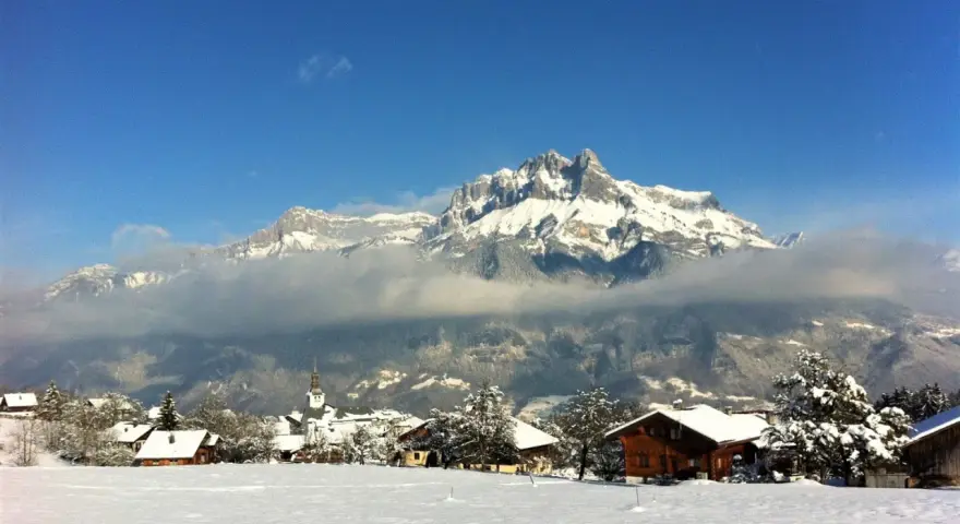 Un pueblo alpino cubierto de nieve, con casas de madera y una iglesia, bajo imponentes montañas nevadas. Un paisaje de invierno en francés.