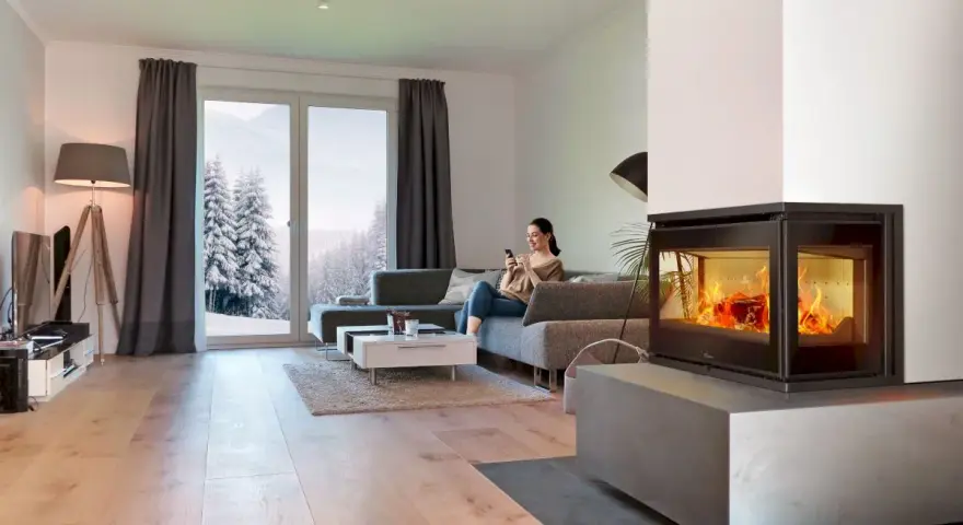 Mujer en sofá mira móvil junto a chimenea de cassette pellets con fuego vivo. Ventana muestra paisaje nevado.