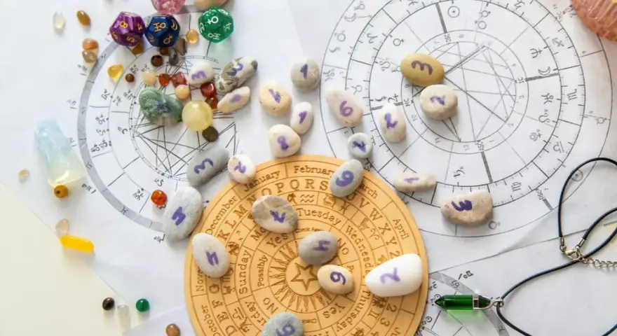 Kamienie z numerami, kostki do gry i talizmany na tle wykresów astrologicznych. Wibracja dnia 5 jest widoczna na jednym z kamieni.