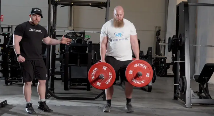 En man med skägg utför farmers walk med tunga vikter. En tränare visar hur övningen ska göras.