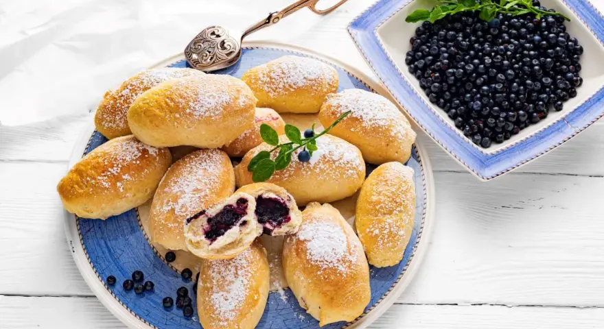 Pyszne jagodzianki posypane cukrem pudrem, obok miseczka pełna świeżych jagód. Idealne na letnie popołudnie.