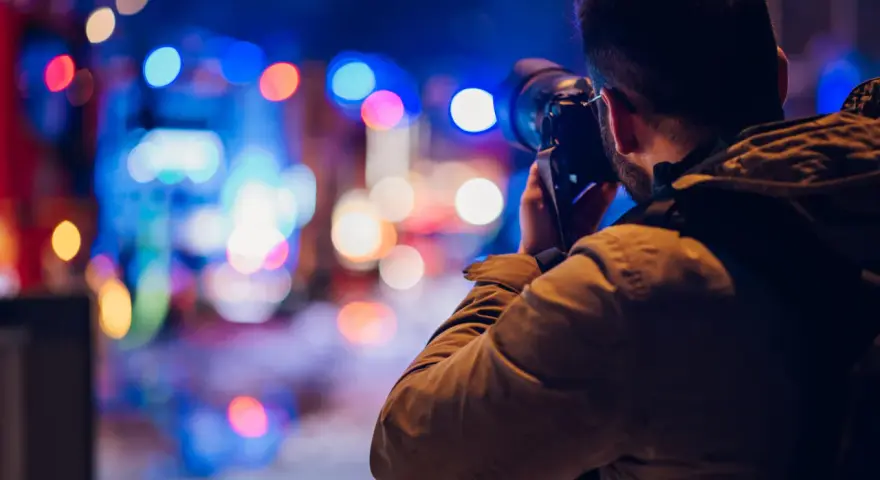Mężczyzna z aparatem w ręku, uchwycony w trakcie nocnej fotografii. Rozmyte, kolorowe światła miasta tworzą magiczne tło.