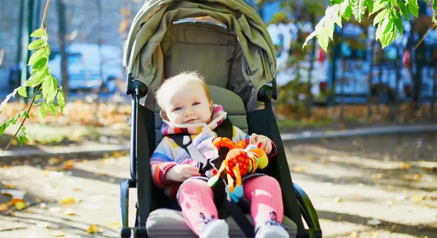Uśmiechnięte dziecko w kolorowym sweterku i różowych skarpetkach siedzi w spacerówce, trzymając zabawkę. Czas na spacer!
