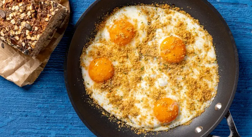 Uova fritte in padella, condite con pangrattato e erbe aromatiche, perfette per le tue ricette con uova. Accanto, una fetta di pane integrale.