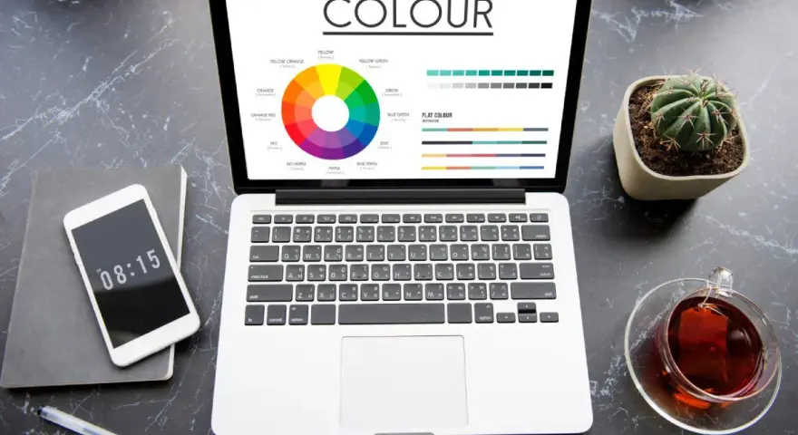 Na ekranie laptopa koło barw i palety kolorów, pomocne przy wyborze między rgb czy cmyk do druku. Obok telefon, notes i kaktus.