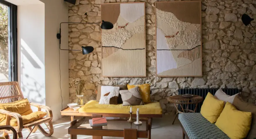 Salon avec mur en pierre, deux tapisseries texturées, fauteuil en rotin, banquette jaune et coussins aux motifs ethniques. Une ambiance deco campagne boheme chaleureuse.