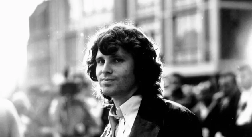 Jim Morrison, mit seinem charakteristischen lockigen Haar und einem nachdenklichen Blick, steht im Gegenlicht.