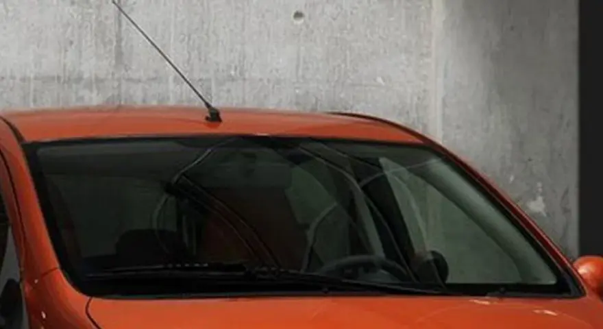 Pomarańczowy samochód z przyciemnianymi szybami z przodu, z antenką na dachu, stoi przy betonowej ścianie.