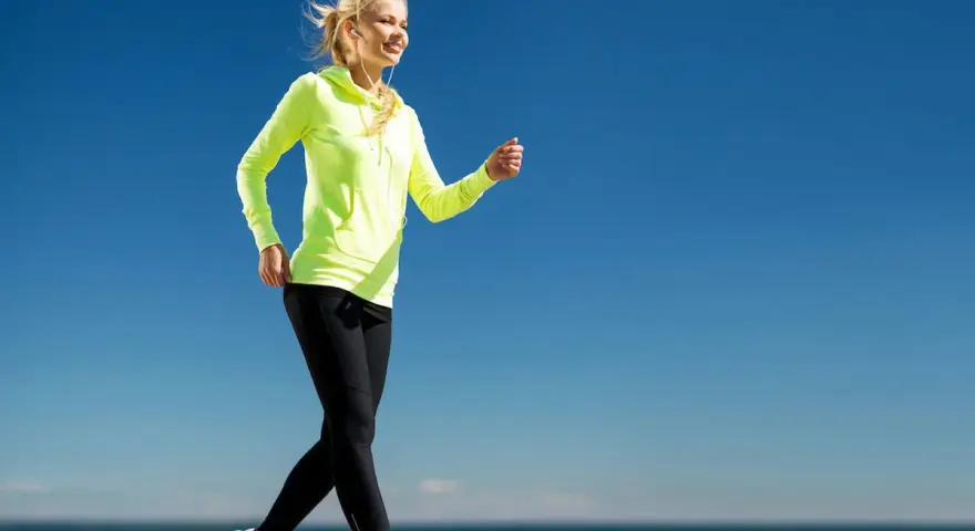 Uśmiechnięta blondynka w neonowej bluzie i czarnych legginsach uprawia power walking nad morzem.