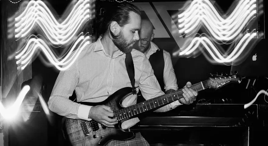 Muzyk gra na gitarze elektrycznej. W tle widać drugiego muzyka i falujące światła, tworzące efekt ekspozycji.
