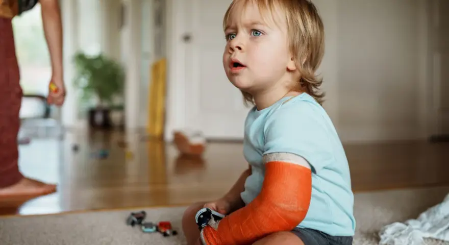 Kleiner Junge mit einem orangefarbenen Gips am Arm, der nachdenklich auf den Boden schaut. Er hat einen **kind arm gebrochen**.