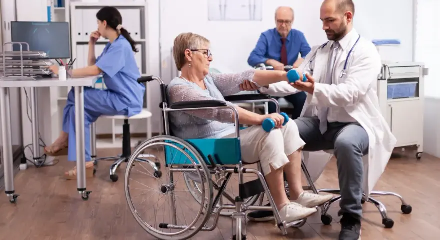 Rehabilitacja po uszkodzeniu móżdżku: pacjentka w wózku wykonuje ćwiczenia z hantlami pod okiem fizjoterapeuty.