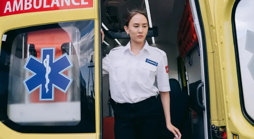 Młoda kobieta w białej koszuli z naszywką "PARAMEDICS" stoi przy otwartych drzwiach karetki. Czy kierowca karetki musi być ratownikiem?