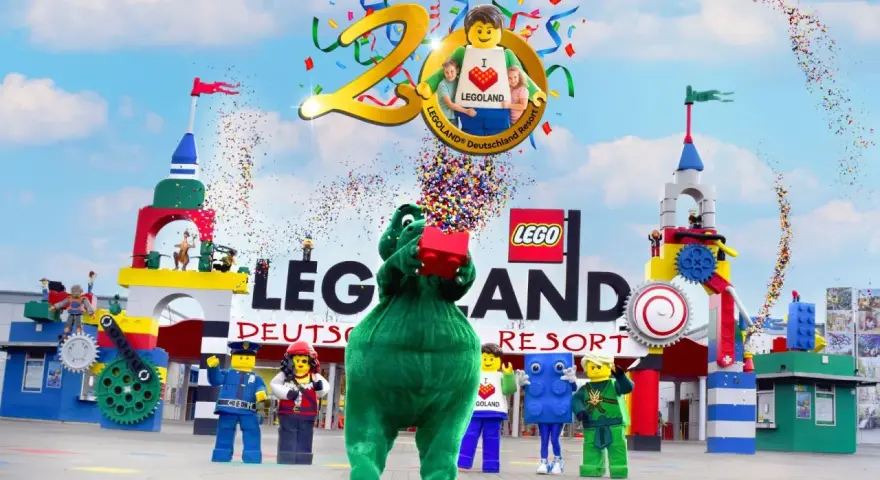 Bunte Konfetti-Explosion bei der Legoland Parade! Ein grüner Drache hält einen roten Lego-Stein hoch, umgeben von Figuren und bunten Türmen.