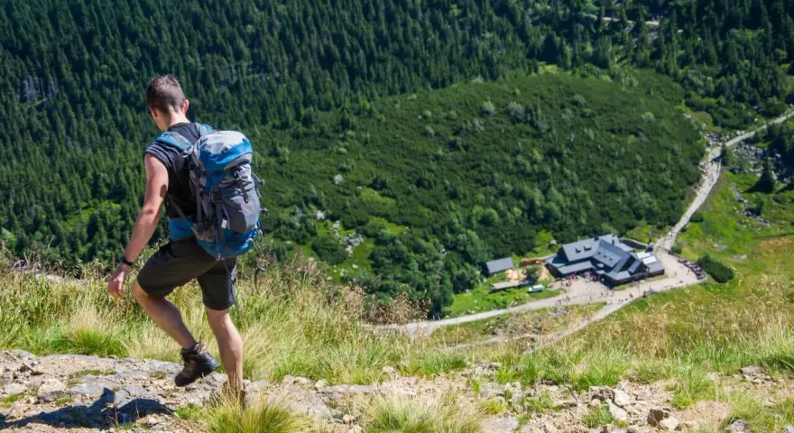 Turysta z plecakiem schodzi po kamienistej ścieżce w Karkonoszach. W dole widoczny schronisko, jedna z wielu atrakcji górskich.
