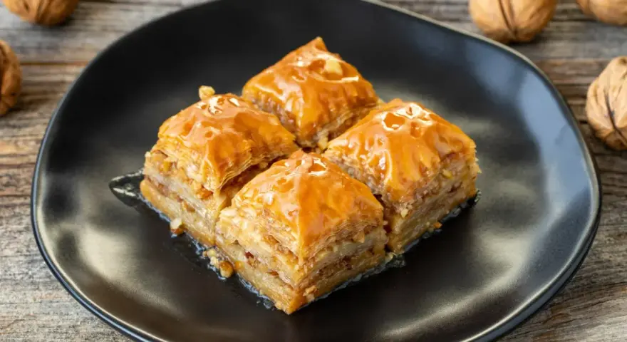 Cztery kawałki baklawy na czarnym talerzu, polane syropem. Obok orzechy włoskie. Idealny baklava przepis na słodki deser.
