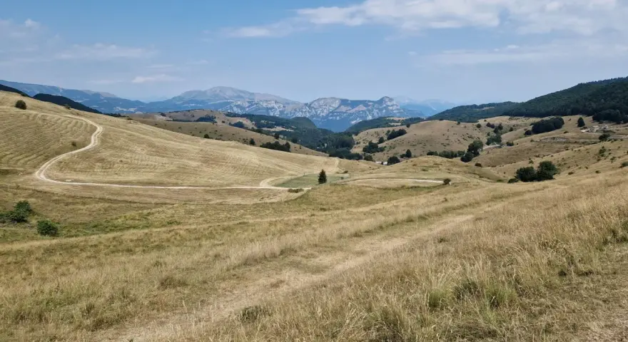 Ampia vallata con erba secca e un sentiero tortuoso che sale verso il passo fittanze. Montagne sullo sfondo sotto un cielo azzurro con nuvole bianche.