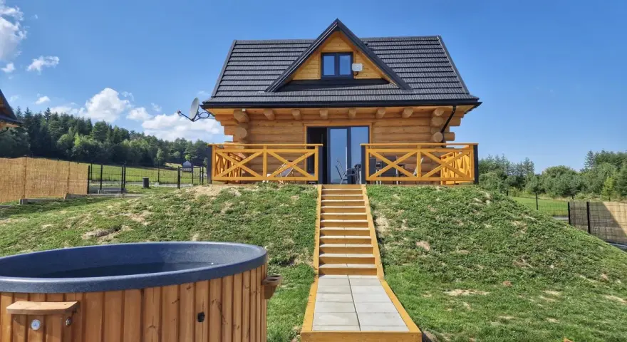 Uroczy domek na odludziu z drewnianą werandą i schodami prowadzącymi do drzwi. Przed domkiem jacuzzi i leżaki.