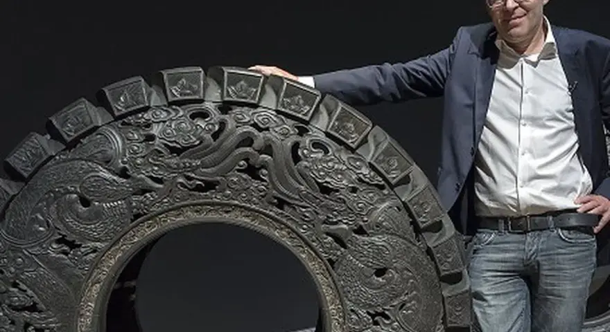 Wim Delvoye pose à côté d'une roue ornée de dragons, une œuvre d'art monumentale.