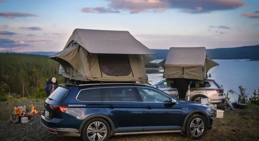 Två bilar med takboxar som fungerar som tält står redo för camping vid en sjö. En eld sprakar i förgrunden. En perfekt plats för camping i Gnesta.