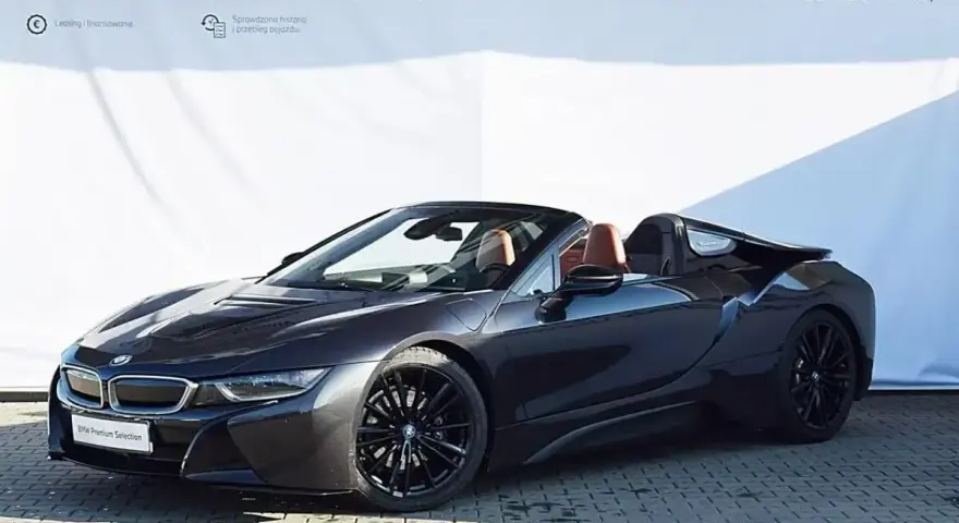 Ciemnoszary kabriolet BMW i8 z brązowym wnętrzem na tle białej ściany z logo BMW.