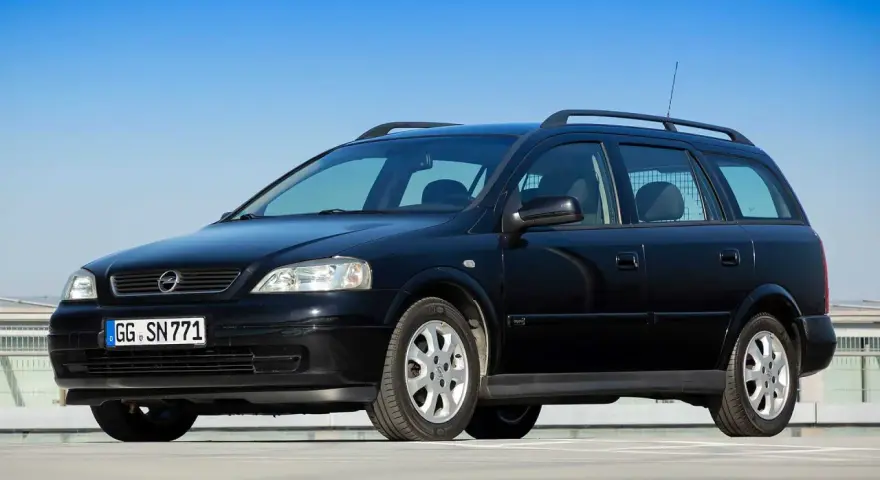 Schwarzer Opel Astra Kombi, alt, mit Dachreling und Kennzeichen GG SN 771, parkt auf einem Parkdeck vor blauem Himmel.
