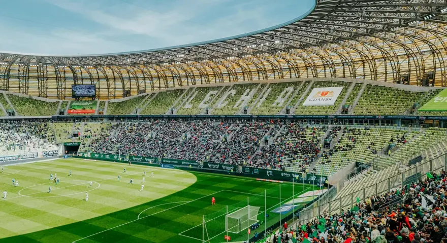 Widok na murawę i trybuny Polsat Plus Arena Gdańsk podczas meczu. Tłumy kibiców na zielonych krzesełkach.