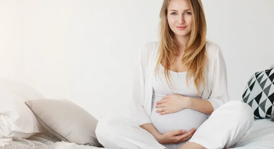 Uśmiechnięta kobieta w ciąży w białych ubraniach siedzi na łóżku, delikatnie trzymając się za brzuch.