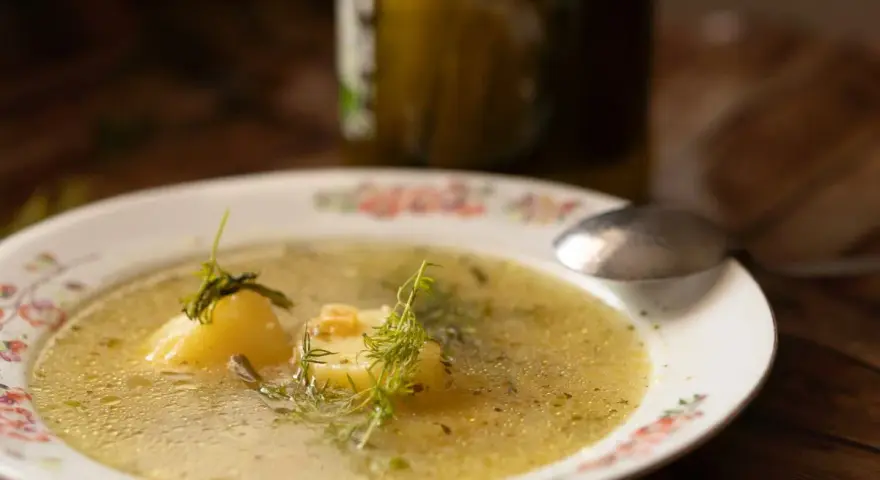 Aromatyczna keto zupa ogórkowa z ziemniakami i koperkiem, podana na talerzu z kwiatowym wzorem. Obok stoi słoik ogórków.
