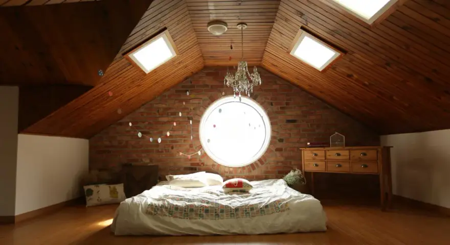 Camera puntata verso un letto in mansarda, con il soffitto in legno e la sua evidente pendenza tetto. Finestre sul tetto e un oblò circolare illuminano la stanza.