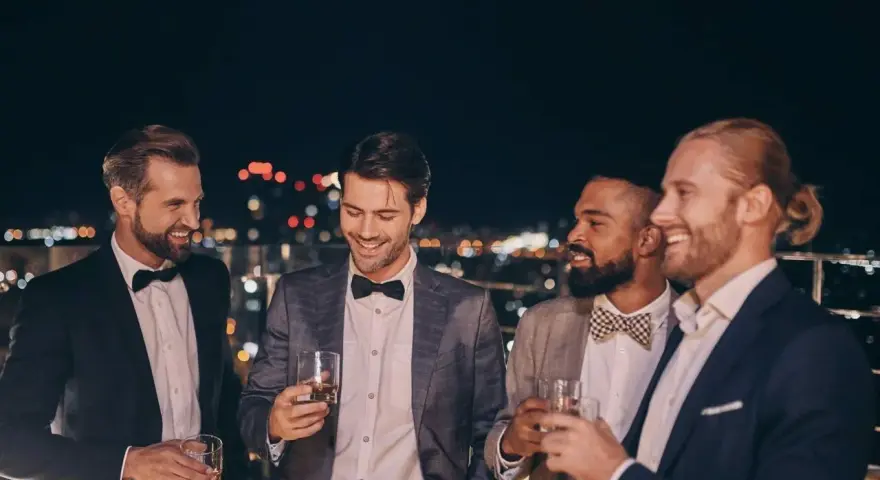Eleganccy panowie w garniturach i muszkach, pijący drinki na tarasie. Idealny przykład, jak się ubrać na poprawiny.