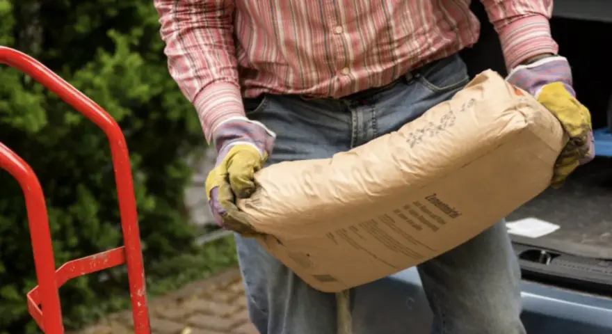Mężczyzna w rękawicach niesie worek cementu. Zastanawia się, ile piasku na 25 kg cementu będzie potrzebne do budowy.
