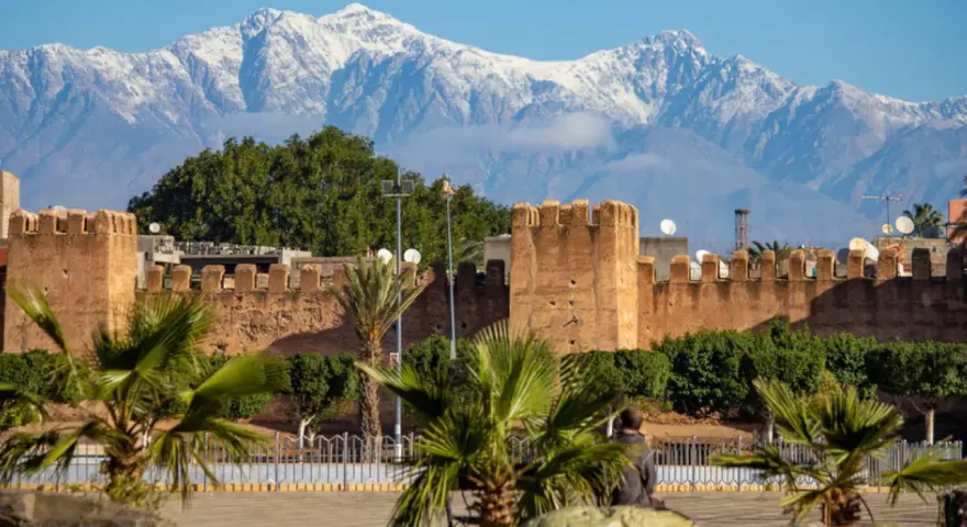 Marrakesz, gdzie leży, widać na tle ośnieżonych gór Atlas. Stare mury miasta i palmy tworzą malowniczy krajobraz.