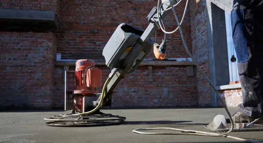 Robot do zacierania betonu pracuje na budowie. Robotnik w niebieskich spodniach kieruje maszyną, która wygładza świeżą posadzkę.