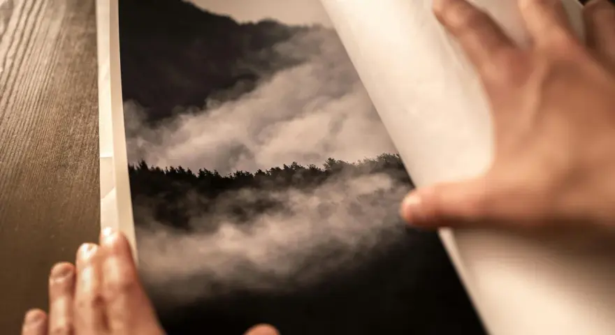 Dłonie otwierają album z fotografią górskiego lasu we mgle, wydrukowaną na papierze barytowym.