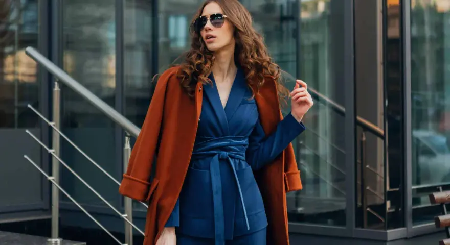 Une femme porte un gilet avec une robe bleu marine, un manteau orange sur les épaules, et des lunettes de soleil.