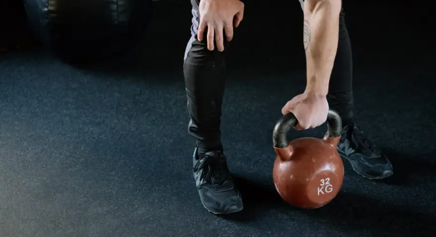 Mężczyzna w czarnych legginsach i butach sportowych podnosi kettlebell 32 kg. Zastanawia się, czy po masażu można iść na trening.