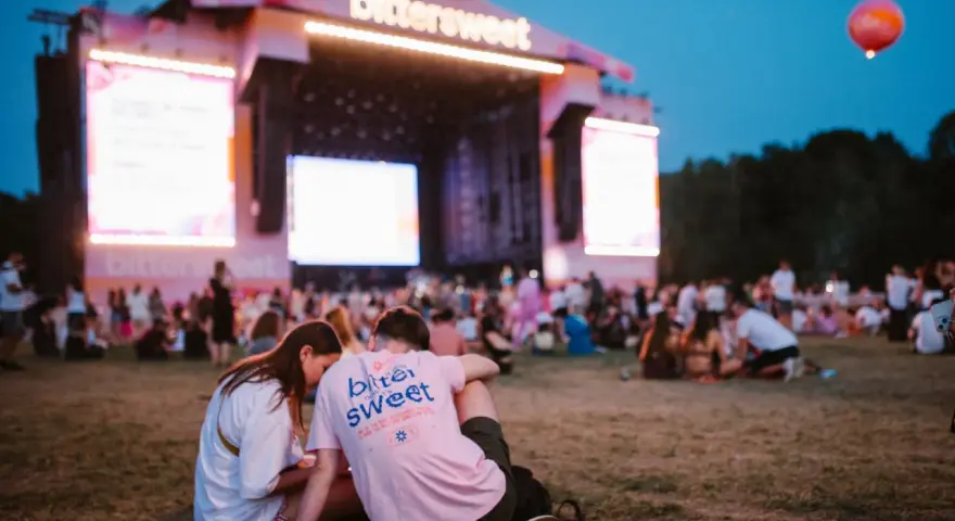 Para siedząca na trawie przed sceną festiwalową z napisem "bittersweet". W tle tłum ludzi i wielkie ekrany.