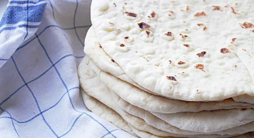 En hög med nybakade tortillabröd, perfekta för ditt nästa tortilla recept. Bredvid ligger en blåvitrutig kökshandduk.