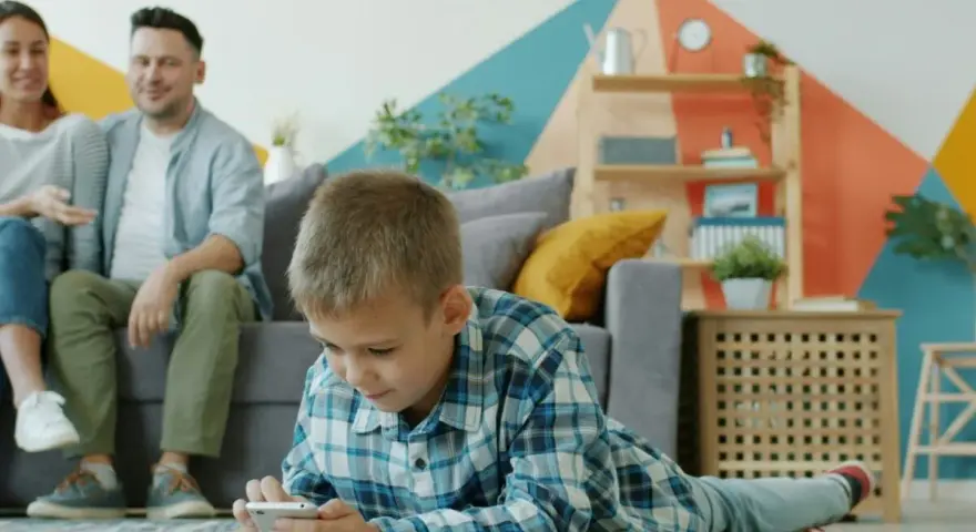 Chłopiec gra na telefonie, podczas gdy rodzice siedzą na kanapie. Ciekawe, od ilu lat jest WhatsApp i czy oni też go używają?
