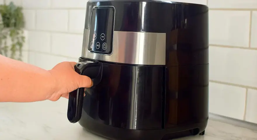 En hand drar ut korgen på en svart airfryer. Den här köksapparaten är lika praktisk som en diskmaskin för snabb matlagning.