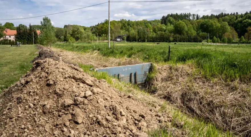Nasyp ziemi przy rowie z niebieską przepustnicą, która może służyć do retencji wody. W tle zielone łąki i las.