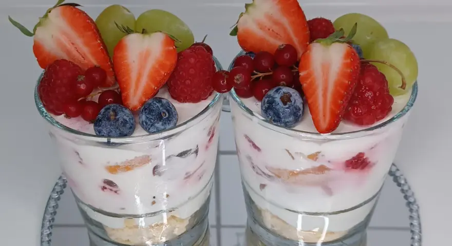 Zwei Gläser mit einem cremigen Dessert, garniert mit frischen Erdbeeren, Himbeeren, Blaubeeren, Johannisbeeren und Trauben. Ein köstliches Dessert im Glas.