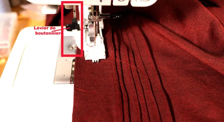 Levier de boutonnière sur machine à coudre, prêt pour réaliser une boutonnière parfaite sur un tissu bordeaux plissé.