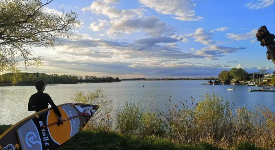 Ein junger Mann mit SUP-Board am Ufer des Alperstedter Sees. Der Himmel ist blau mit weißen Wolken, die Sonne scheint.