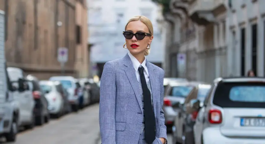 Mujer con traje a cuadros, camisa blanca y corbata negra, luciendo un impecable tomboy style. Gafas de sol y labios rojos completan su look.