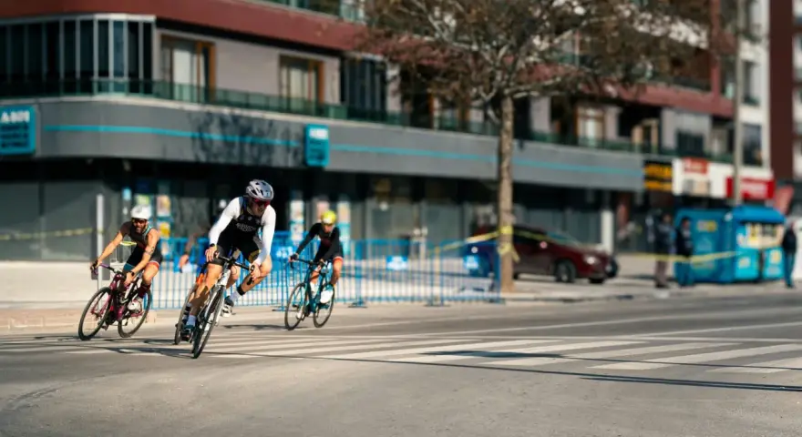 Cyclists race through a city street, perhaps part of le grand tour de France, with buildings and spectators lining the course.